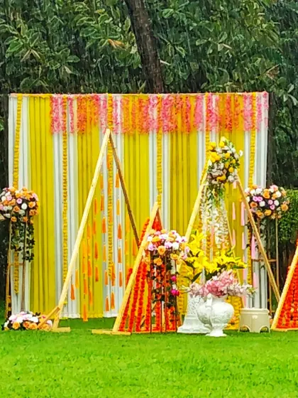 A modern and geometric Haldi decor setup in a garden, using triangular wooden frames and marigold decorations against a yellow and white striped backdrop.
