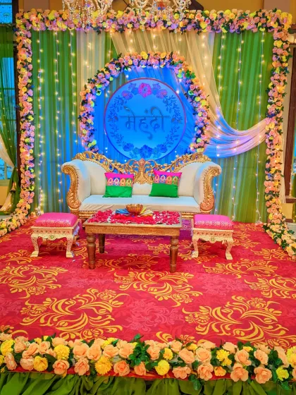 A festive Mehndi stage setup with a comfortable ornate sofa, a custom "Mehndi" sign, and a backdrop of green drapes and circular floral arrangements lit with fairy lights.