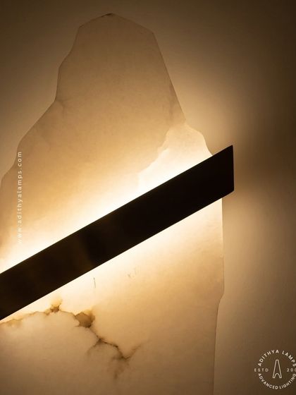 This artistic wall sconce features a raw, organically shaped slab of translucent marble. A simple diagonal metal bar houses the light, which glows through the stone to reveal its natural veining and character.