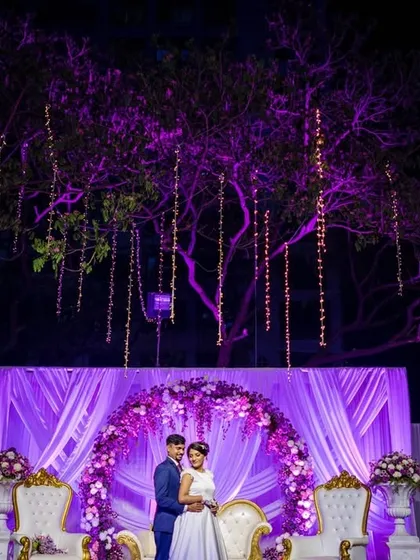 A grand reception stage with the couple framed by an arch of purple flowers and hanging lights. This portrait showcases the beautiful decor and scale of the event.