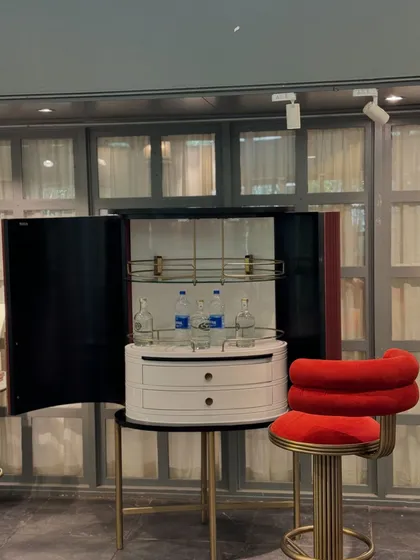 Our compact, elegant bar cabinet on display. This piece features a curved design, a high-gloss finish, and a fully-fitted interior, paired with a vibrant red velvet bar stool.
