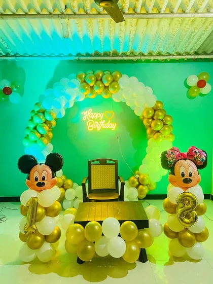 A classic Mickey and Minnie Mouse themed birthday decoration. The setup features a gold and white circular balloon arch and character balloon sculptures holding foil numbers.