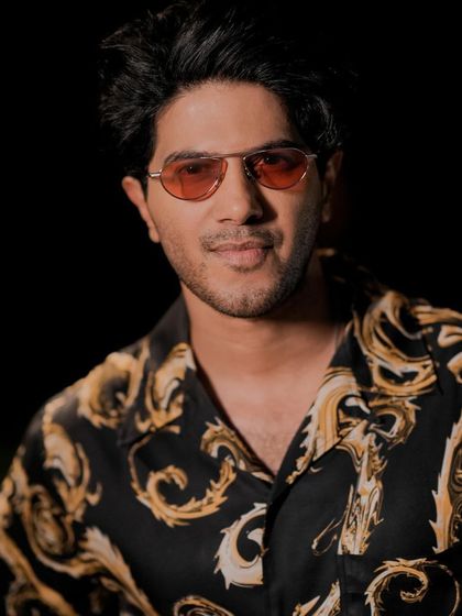 A close-up of Dulquer. The orange-tinted sunglasses pick up the gold tones in the shirt, tying the whole look together.