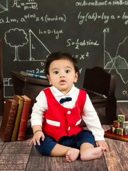 A baby boy in a red vest and bow tie sits in front of his chalkboard, ready for his lesson.