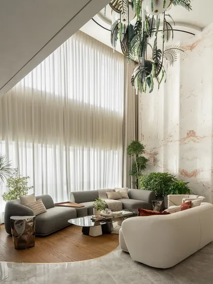The sheer scale of this double-height living room is balanced by the warmth of natural materials and lush indoor plants. The custom chandelier adds a sculptural element, while the mix of wood and marble flooring defines the circular seating area.