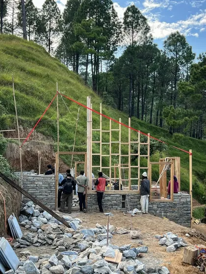 The wooden skeleton of a guesthouse rises from its stone foundation in Uttarakhand. This image captures the initial phase of framing, where the building's form and relationship to the sloping site are first established.