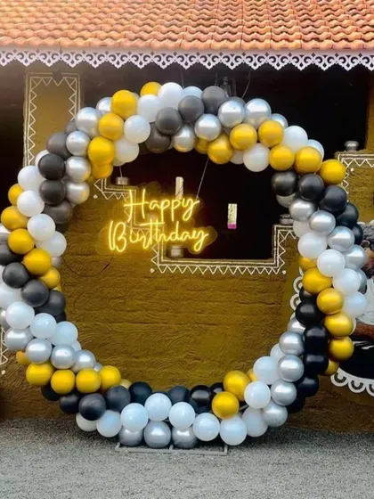 A stylish outdoor birthday setup with a circular balloon arch in black, white, yellow, and silver. The neon 'Happy Birthday' sign adds a modern touch to the rustic background.