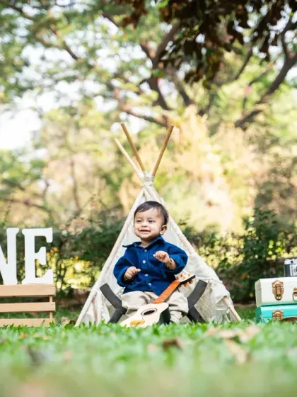 A beautifully styled outdoor first birthday photoshoot featuring a baby boy sitting in front of a teepee, surrounded by props like a 'ONE' sign, vintage suitcases, and a globe.