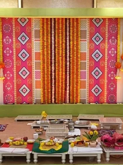 A colorful stage setup for a pre-wedding puja. The backdrop uses printed fabric with traditional motifs and is framed by marigold garlands, creating a vibrant and auspicious setting.