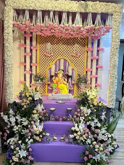 A majestic purple-themed Ganpati mandap. The design features a traditional golden arch backdrop, framed by columns of white and pink flowers and a base filled with lush floral arrangements.