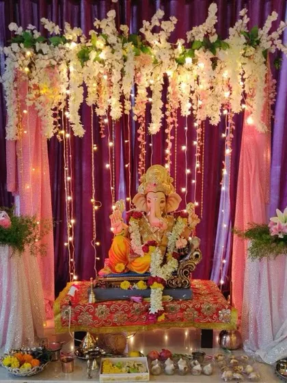A dreamy Ganpati mandap created with a canopy of white flowers and cascading fairy lights against a purple curtain. The sequined fabric on the side tables adds a soft, celebratory shimmer.