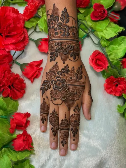 A beautiful party henna design with a floral cuff and delicate finger details, set against a backdrop of red flowers.