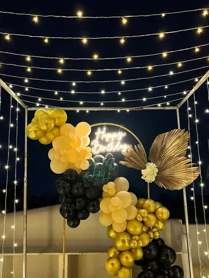 A magical outdoor terrace setup featuring a canopy of fairy lights over a black and gold balloon backdrop with a neon sign.
