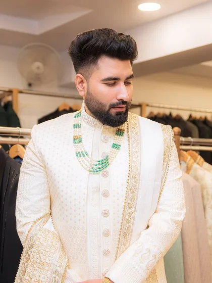 A close-up of a groom-to-be in an elegant ivory sherwani, accessorized with a beautiful green and pearl mala and a gold-trimmed stole.