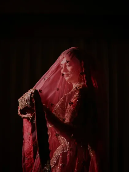 A candid, emotional moment of the bride adjusting her veil. The soft, dramatic lighting creates a silhouette effect, highlighting her smile and the delicate fabric of her dupatta.