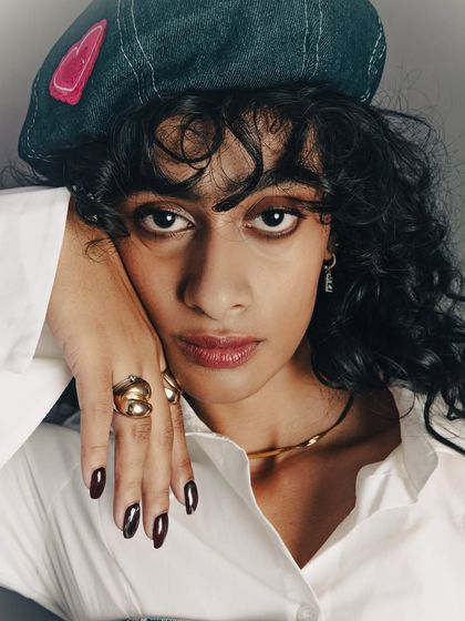 This close-up shot highlights multiple accessories at once: the rings, necklace, and nail polish. The model's hand is posed elegantly to display the jewelry naturally. The lighting is soft yet directional, bringing out the shine of the gold and the deep color of the nails.