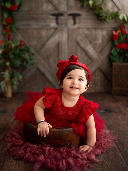 A sweet baby girl in a beautiful red dress, set against a rustic barn wood background.