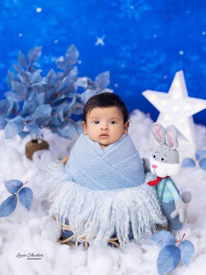 This alert newborn is wrapped snugly in a blue swaddle, looking curious amidst a winter-themed set with a glowing star and a knitted bunny prop.