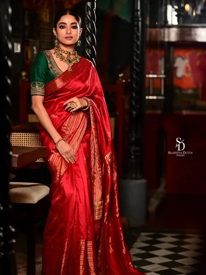 A classic, elegant pose by Ishaa Saha in the red saree. This full-length portrait showcases the beautiful drape and sheen of the fabric in a sophisticated indoor setting.