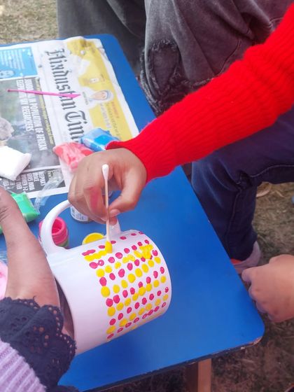 I guide a child as she carefully applies paint to her mug using a cotton bud. This technique is great for creating dot patterns and is easy for small hands to manage.