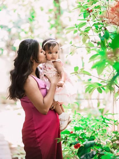 A mother gives her baby a gentle kiss on the cheek amidst lush greenery. This image, from a butterfly-themed birthday shoot, is tender and full of love.