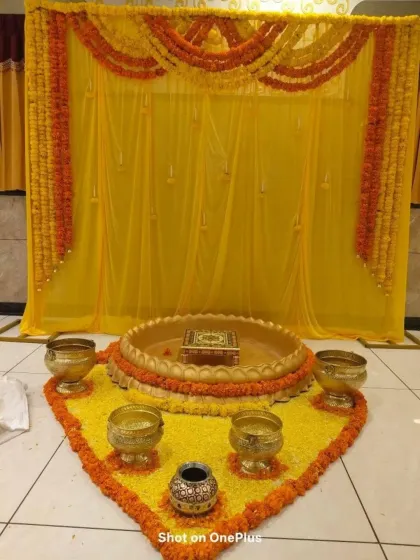 A simple and traditional Haldi ritual area. The space is defined by a yellow floral carpet, brass bowls, and a backdrop of yellow drapes and marigold garlands.