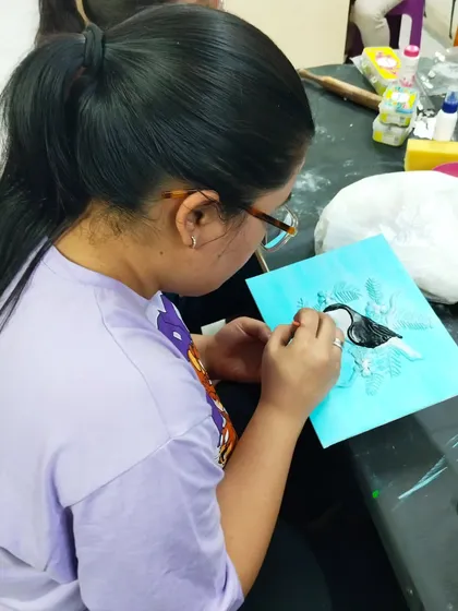 A student painting a bird design on a blue background, fully absorbed in the creative process during a craft workshop.