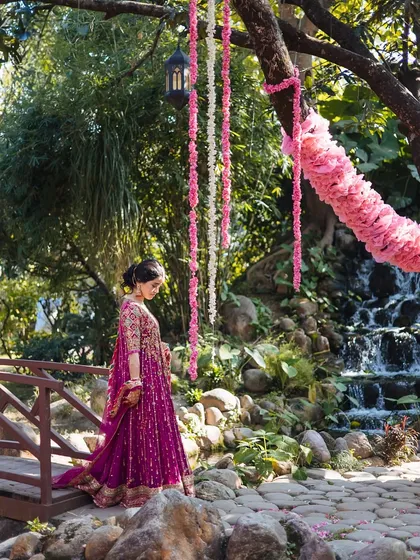 A bride stands on a rustic bridge, her magenta Anarkali a beautiful pop of color against the natural backdrop.