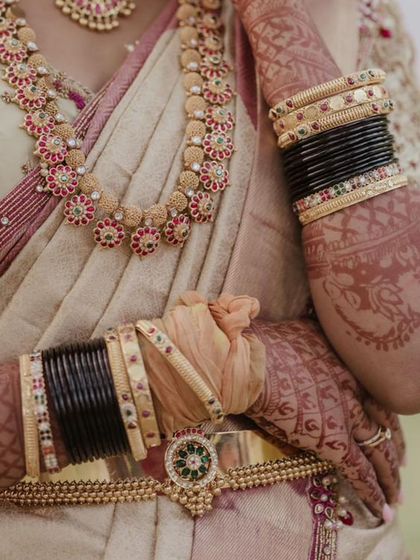 A detail shot focusing on the bride's hands, adorned with henna and traditional bangles, against the rich fabric of her saree. The gold belt adds a classic touch to her waist.