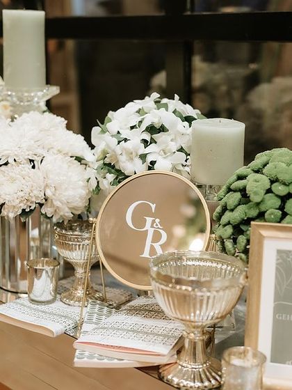 A beautifully styled welcome table featuring personalized signage, framed photos, and elegant glassware. The arrangements of white chrysanthemums and green button mums add a touch of fresh, modern elegance.