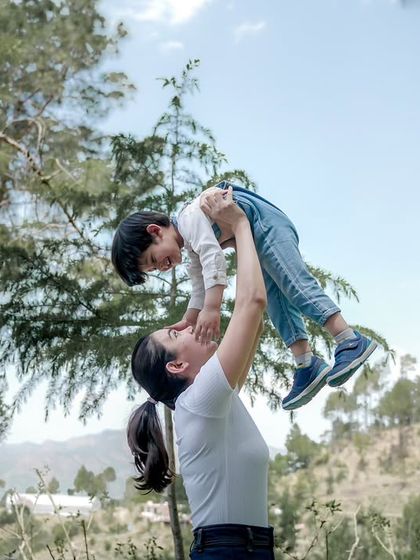 A mother playfully lifts her son into the air against a bright blue sky and green trees.