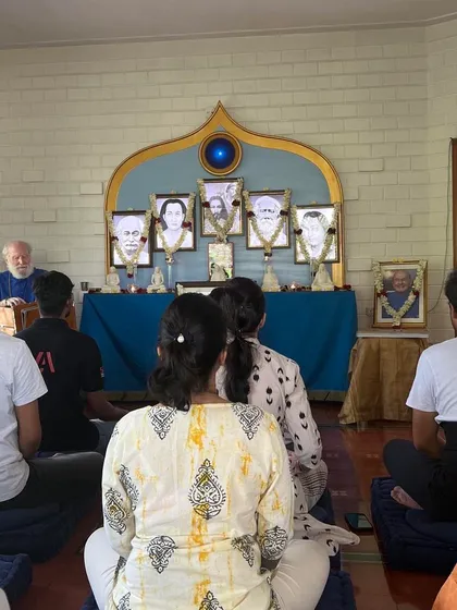 A view from the back of the meditation hall, showing devotees facing the altar. This perspective highlights the shared focus and reverence of the group as they listen to a spiritual discourse.