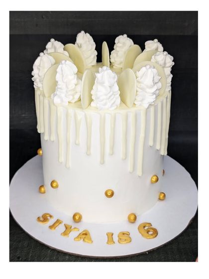 A tall, all-white cake for a white-themed birthday party. This choco-chip cake features a white chocolate drip and is accented with gold spheres for a touch of elegance.