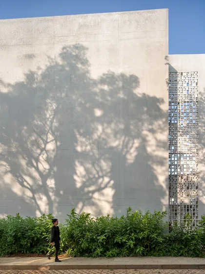The shadow of a large tree is cast upon the textured wall of Vidyashilp Academy, next to a vertical strip of 'jaali'. This image highlights our commitment to integrating the natural landscape, preserving mature trees and making them part of the architectural experience.