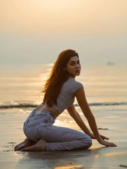 A low-angle shot on the wet sand, reflecting the soft colors of the sunset. The pose is both relaxed and elegant.