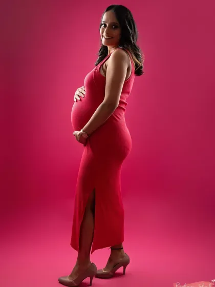 A full-length portrait showcasing a vibrant red dress against a matching pink backdrop. This side profile highlights the baby bump beautifully, creating a bold and confident maternity photo.