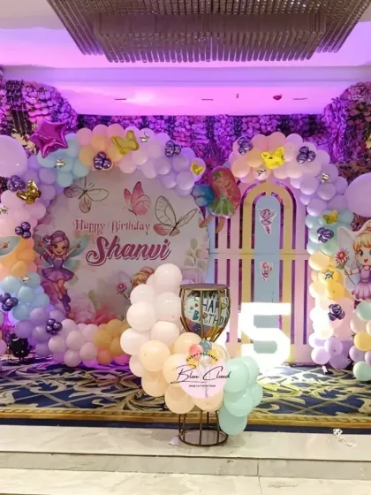 A beautiful fairy-themed stage decoration for a 5th birthday, with pastel balloons, character cutouts, and a light-up number.