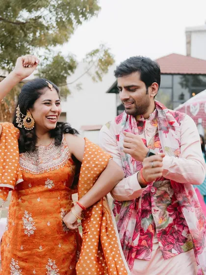 Pure happiness at a sundowner mehendi. The couple is dancing, and their coordinated floral and pastel outfits look fantastic. The groom's floral jacket and the bride's orange sharara are a perfect match.