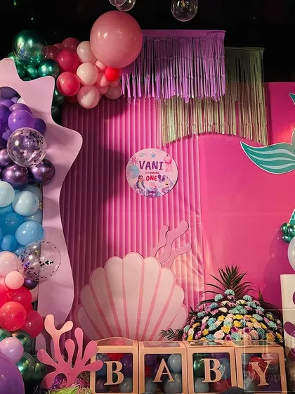 A complete view of Vani's mermaid birthday party stage. The vibrant setup includes a pink textured backdrop, a large seashell prop, coral cutouts, and an explosion of balloons to create an under-the-sea wonderland.