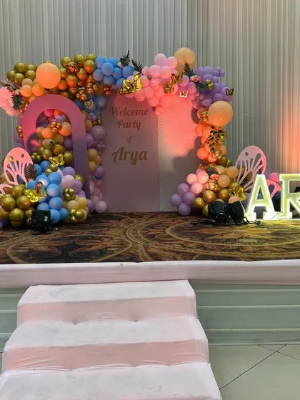 A vibrant butterfly-themed stage for Arya's welcome party. The setup includes a colorful balloon arch, butterfly props, and illuminated marquee letters spelling out her name.