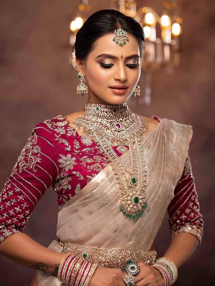 A stunning portrait of a bride in a silver and magenta saree with layered diamond necklaces. The makeup is glamorous, with defined eyes and a flawless base to complement the sparkle of her jewelry.