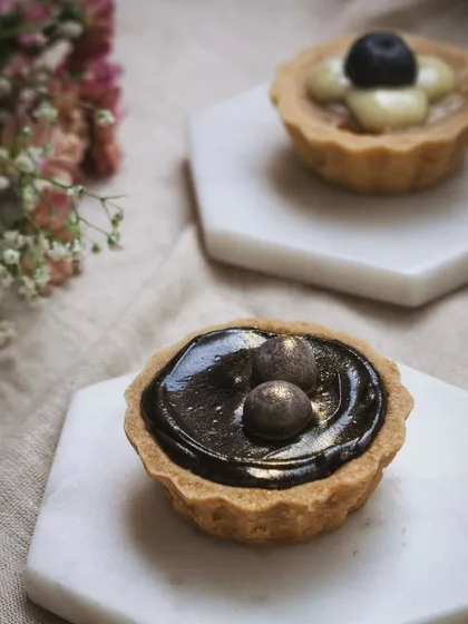 A pair of gourmet tarts, one chocolate and one fruit, on minimalist marble coasters. The soft lighting and simple styling emphasize the fine details and artistry of the pastries.