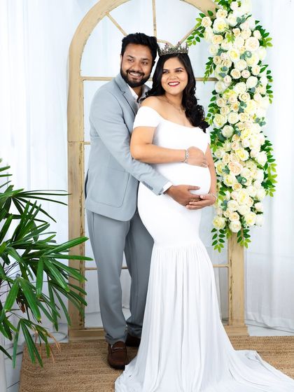 A classic and elegant couple's portrait. The mom-to-be in a white gown and her partner in a grey suit stand together in our beautiful floral archway studio setup.