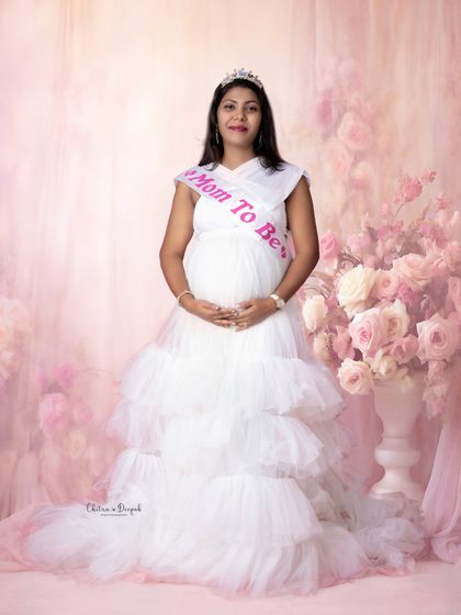 A baby shower theme! This mom-to-be looks radiant in a white gown and a "Mom To Be" sash, surrounded by a soft pink floral backdrop.