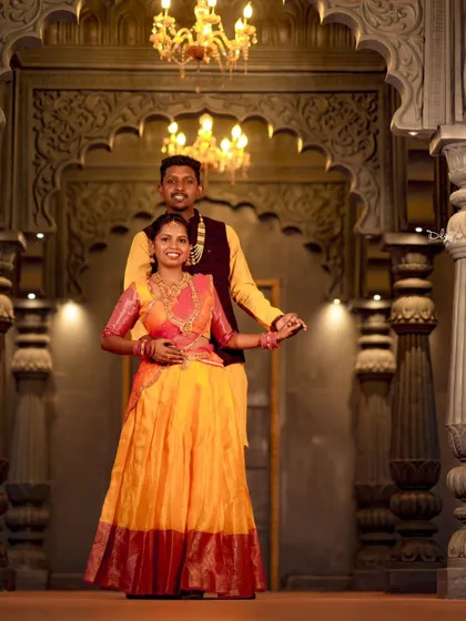 A full-length view of the couple's stunning traditional attire inside a palace-like setting. The bride's orange and pink lehenga is particularly eye-catching.