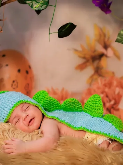 A sleepy newborn dressed as a little dinosaur, nestled in a cozy fur-lined basket. The hand-knitted costume adds a touch of handmade charm.