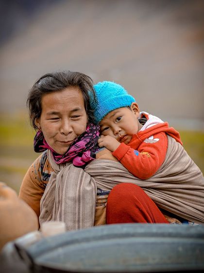 A tender moment between a mother and child in Himachal Pradesh. The child, wrapped warmly on her back, looks towards the camera with innocent eyes.