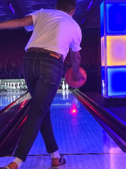A classic bowling shot from behind the player, showing the ball rolling towards the pins under the cool blue and yellow lights.