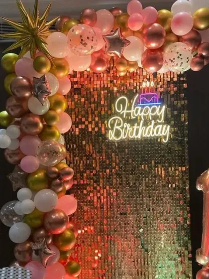 A birthday party with a rose gold shimmer wall, a 'Happy Birthday' neon sign, and a balloon garland.