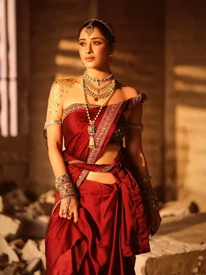 A full-length shot of Nyra Banerjee in a rich, red saree, styled with a modern drape to showcase movement. The look is completed with layered necklaces and arm cuffs, set against a rustic, sun-lit background.